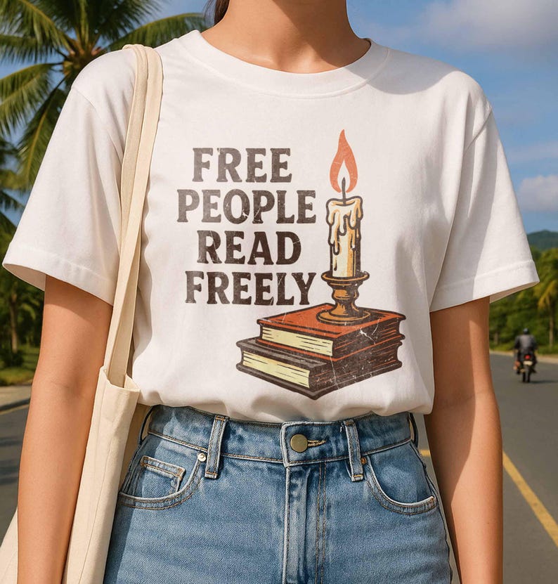 Może przedstawiać: Biała koszulka z napisem "FREE PEOPLE READ FREELY" w brązowych literach. Koszulka przedstawia grafikę świecy na dw&oacute;ch książkach. Świeca ma ż&oacute;łty płomień i jest w złotym uchwycie.