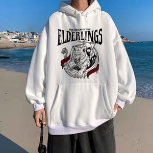 May include: White hooded sweatshirt with a front pocket and a graphic design. The graphic features a wolf, a Viking ship, and the text "THE REALM OF THE ELDERLINGS" in black. The sweatshirt has white cuffs and a white bottom hem.