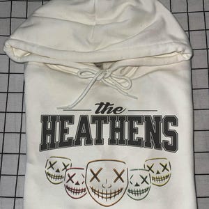 May include: Cream-coloured hoodie featuring the text "The Heathens" in dark grey. Below are five smiling mask graphics with "X" eyes. The hoodie also has the quote, "EVEN IF YOU HATE YOURSELF. I'LL LOVE YOU FOR THE BOTH OF US."