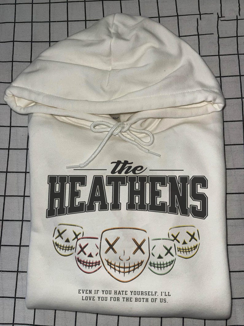 May include: Cream-coloured hoodie featuring the text "The Heathens" in dark grey. Below are five smiling mask graphics with "X" eyes. The hoodie also has the quote, "EVEN IF YOU HATE YOURSELF. I'LL LOVE YOU FOR THE BOTH OF US."