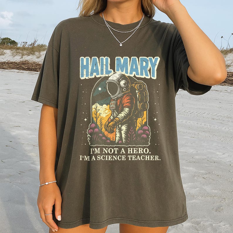 May include: Dark olive green t-shirt featuring a graphic of an astronaut with the text "HAIL MARY" above and "I'M NOT A HERO. I'M A SCIENCE TEACHER." below. The astronaut is in a spacesuit with a backpack, set against a space and plant background.