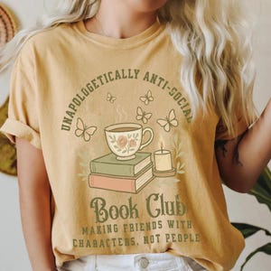 May include: Mustard yellow t-shirt with a graphic design. The design features a teacup with roses, books, a candle, and butterflies. Text reads "UNAPOLOGETICALLY ANTI-SOCIAL BOOK CLUB MAKING FRIENDS WITH CHARACTERS, NOT PEOPLE."