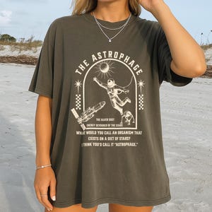 May include: Dark gray t-shirt with a graphic design featuring an astronaut, a sun, and the text "THE ASTROPHAGE". The design includes the text "THE SILVER DUST ENERGY DEVOURER OF THE STARS".
