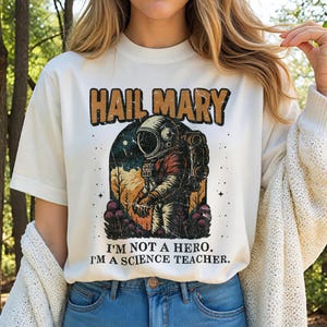 May include: Cream-colored t-shirt with the text "HAIL MARY" in brown and orange lettering. Below the text is an illustration of an astronaut in a spacesuit. The text "I'M NOT A HERO. I'M A SCIENCE TEACHER." is printed below the illustration.
