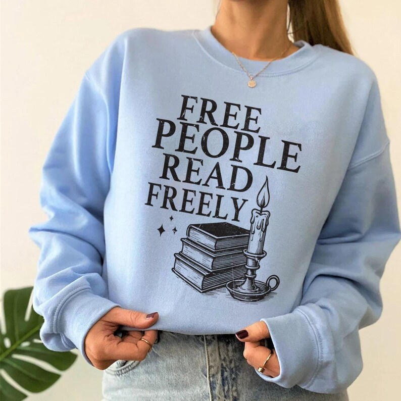 Op de afbeelding: Een lichtblauwe sweatshirt met de zwarte tekst "FREE PEOPLE READ FREELY". De afbeelding bevat een stapel boeken, een brandende kaars en sterren. De sweatshirt heeft lange mouwen en een ronde hals.
