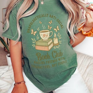 May include: Green t-shirt with the text "UNAPOLOGETICALLY ANTI-SOCIAL" and "Book Club". The graphic features a teacup, books, butterflies, and a candle. The shirt is a comfortable, casual style.