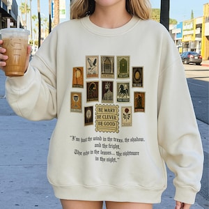 May include: Cream-colored sweatshirt featuring a collection of vintage-style postage stamps with various illustrations and text. Below the stamps is the phrase "BE WARY BE CLEVER BE GOOD" and a quote. A beverage in a clear cup is held in the left hand.
