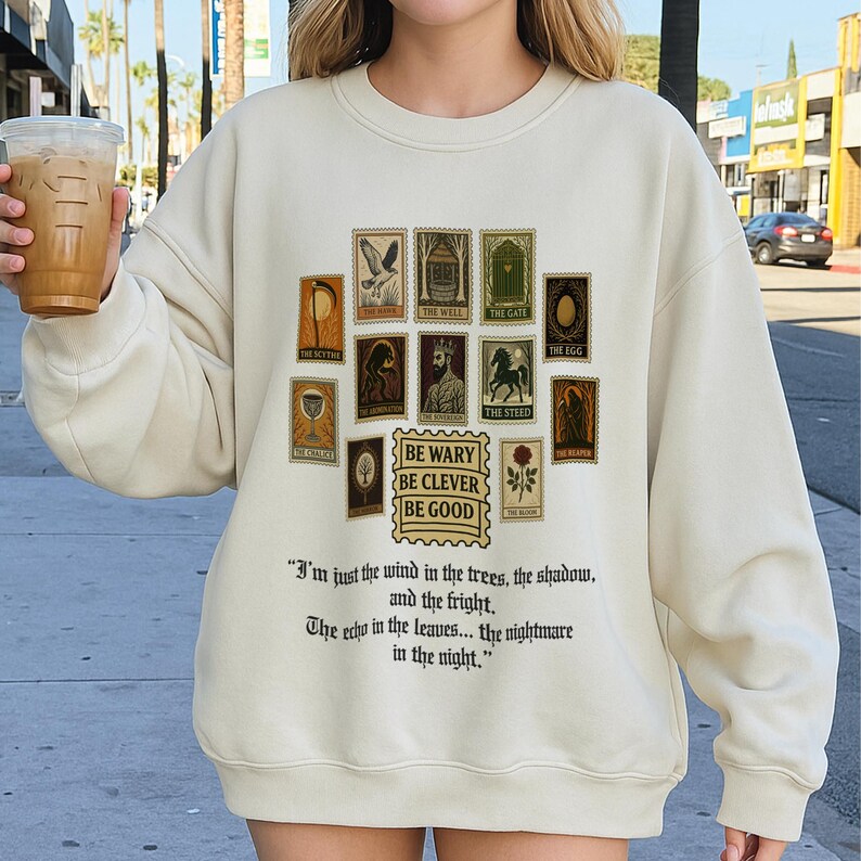 May include: Cream-colored sweatshirt featuring a collection of vintage-style postage stamps with various illustrations and text. Below the stamps is the phrase "BE WARY BE CLEVER BE GOOD" and a quote. A beverage in a clear cup is held in the left hand.