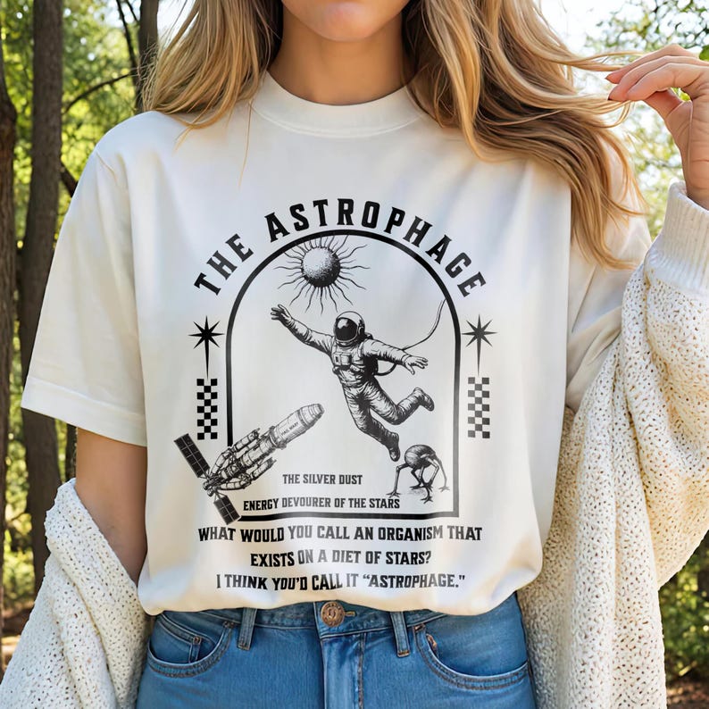 May include: Cream-colored t-shirt with a black graphic design featuring an astronaut, sun, and spacecraft. Text reads "THE ASTROPHAGE" and "THE SILVER DUST, ENERGY DEVOURER OF THE STARS." Additional text asks, "WHAT WOULD YOU CALL AN ORGANISM THAT EXISTS ON A DIET OF STARS? I THINK YOU'D CALL IT 'ASTROPHAGE.'"