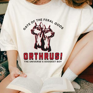 Puede incluir: Camiseta color crema con un gr&aacute;fico de un perro de dos cabezas, el texto "GATE OF THE FERAL GODS" arriba y "ORTHRUS! THE UNIVERSE'S GOODEST BOY" abajo. La camiseta est&aacute; hecha de un material suave.