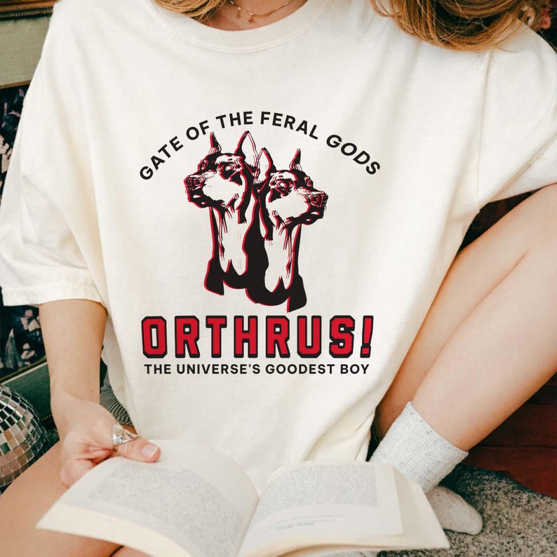 Puede incluir: Camiseta color crema con un gr&aacute;fico de un perro de dos cabezas, el texto "GATE OF THE FERAL GODS" arriba y "ORTHRUS! THE UNIVERSE'S GOODEST BOY" abajo. La camiseta est&aacute; hecha de un material suave.