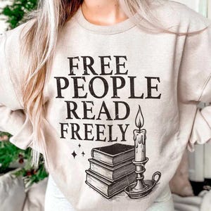 Op de afbeelding: Beige sweatshirt met de zwarte tekst "FREE PEOPLE READ FREELY". Onder de tekst staat een afbeelding van drie gestapelde boeken, een kaars en een kandelaar. Twee kleine ruitvormen staan links van de boeken.