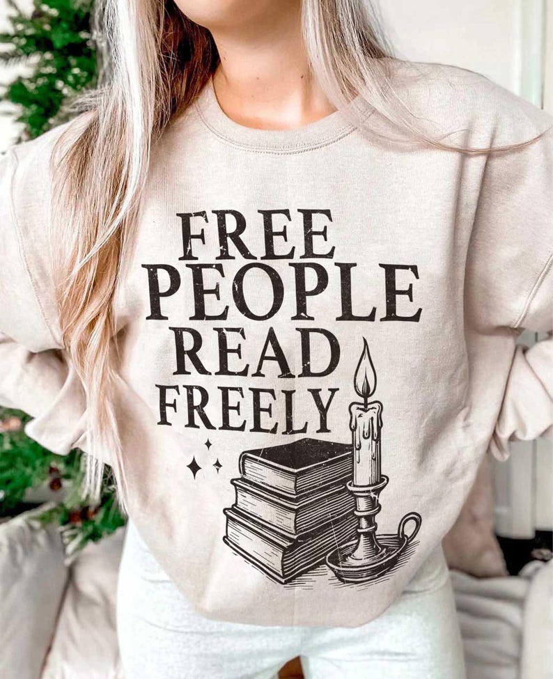 Op de afbeelding: Beige sweatshirt met de zwarte tekst "FREE PEOPLE READ FREELY". Onder de tekst staat een afbeelding van drie gestapelde boeken, een kaars en een kandelaar. Twee kleine ruitvormen staan links van de boeken.