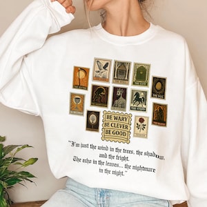May include: White sweatshirt featuring a collection of vintage-style postage stamps with various illustrations and text. The stamps include images of a scythe, a hawk, a well, a gate, an egg, and more. The text "BE WARY BE CLEVER BE GOOD" is also present.