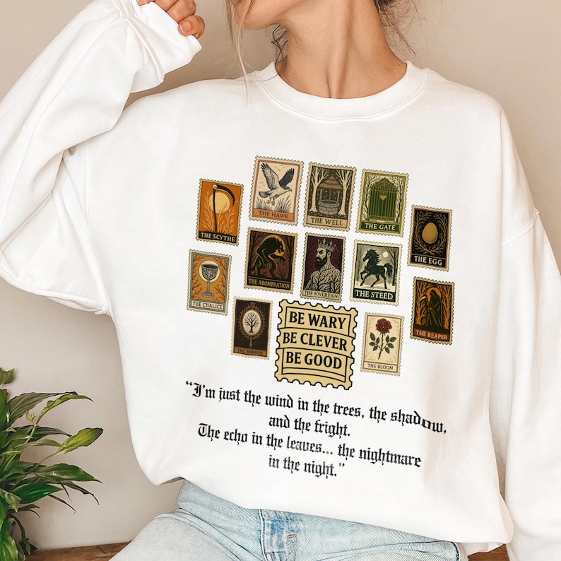 May include: White sweatshirt featuring a collection of vintage-style postage stamps with various illustrations and text. The stamps include images of a scythe, a hawk, a well, a gate, an egg, and more. The text "BE WARY BE CLEVER BE GOOD" is also present.