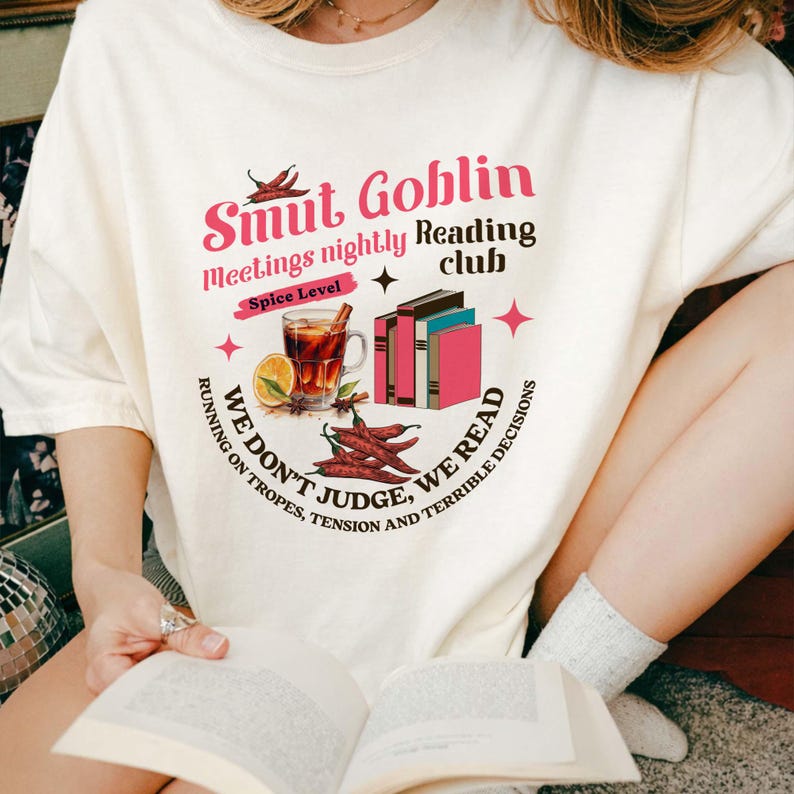 May include: Cream-colored t-shirt with a graphic design featuring the text "Smut Goblin Reading Club." The design includes illustrations of books, a spiced drink, and chili peppers. The shirt also has the phrases "Meetings nightly," "Spice Level," and "We don't judge, we read."