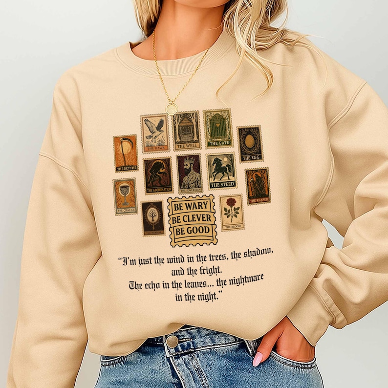 May include: Beige sweatshirt featuring a collection of postage stamp-style graphics with various images and text. The text "BE WARY BE CLEVER BE GOOD" is also present. Below the graphics is a quote about the wind and the night.