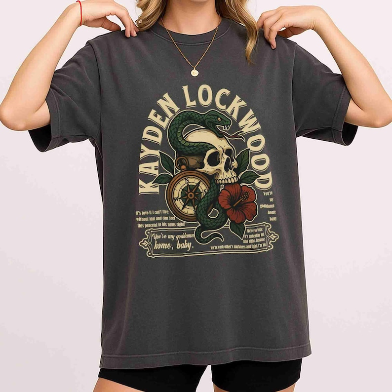 May include: Dark gray t-shirt featuring a graphic design with the text "KAYDEN LOCKWOOD". The design includes a skull, snake, compass, and a red hibiscus flower. The shirt has short sleeves and a crew neck.