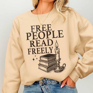 Op de afbeelding: Beige sweatshirt met de zwarte tekst "FREE PEOPLE READ FREELY", samen met een afbeelding van boeken, een kaars en sterren. De trui heeft een ronde hals en lange mouwen.