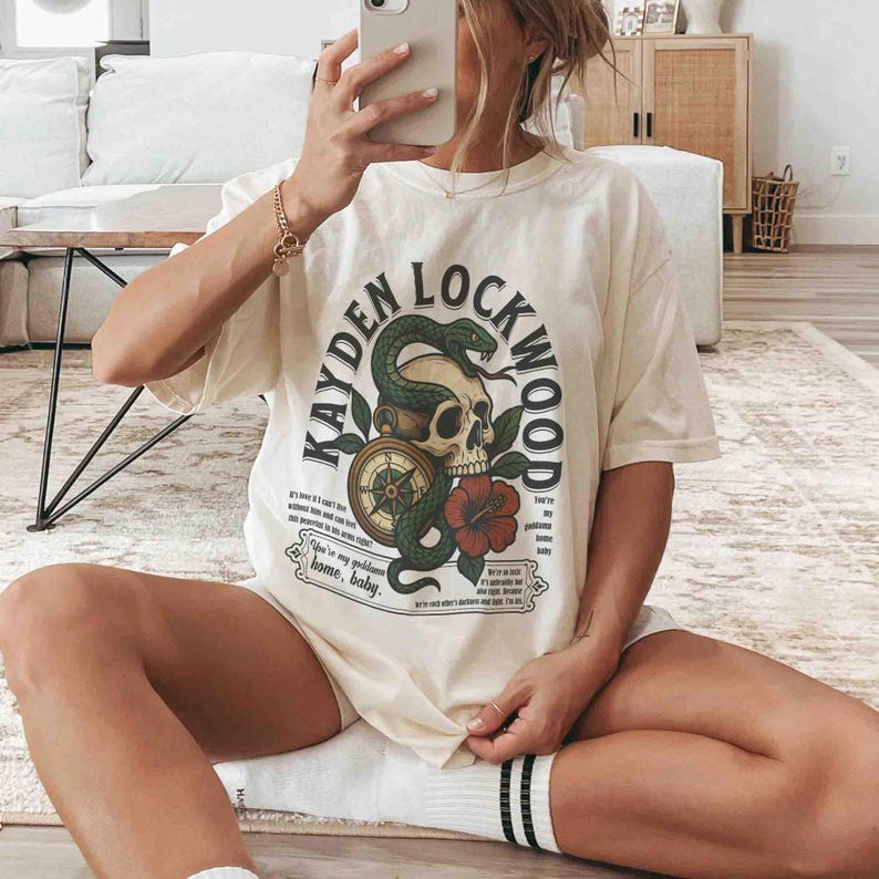 May include: Cream-colored t-shirt featuring a graphic design with the text "KAYDEN LOCKWOOD". The design includes a skull, a snake, a compass, and a red flower. The shirt has short sleeves and a relaxed fit.