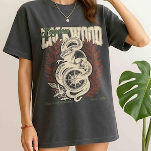 Puede incluir: Camiseta gris oscuro con un dise&ntilde;o gr&aacute;fico desgastado que presenta una calavera, una serpiente y una br&uacute;jula. El texto "Kayden Lockwood" est&aacute; encima del gr&aacute;fico, con la frase "You're my goddamn home, baby" debajo.