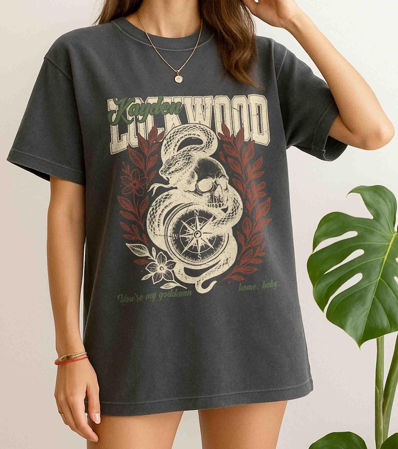 Puede incluir: Camiseta gris oscuro con un dise&ntilde;o gr&aacute;fico desgastado que presenta una calavera, una serpiente y una br&uacute;jula. El texto "Kayden Lockwood" est&aacute; encima del gr&aacute;fico, con la frase "You're my goddamn home, baby" debajo.