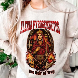 Op de afbeelding: Beige sweatshirt met een afbeelding van een vrouw in een rode mantel en kroon, met een kruis. De tekst "ALEXIA PYROGENNETOS" staat in het rood boven de afbeelding, met "The Heir of Troy" eronder. Vlammen en een schedel zijn ook aanwezig.