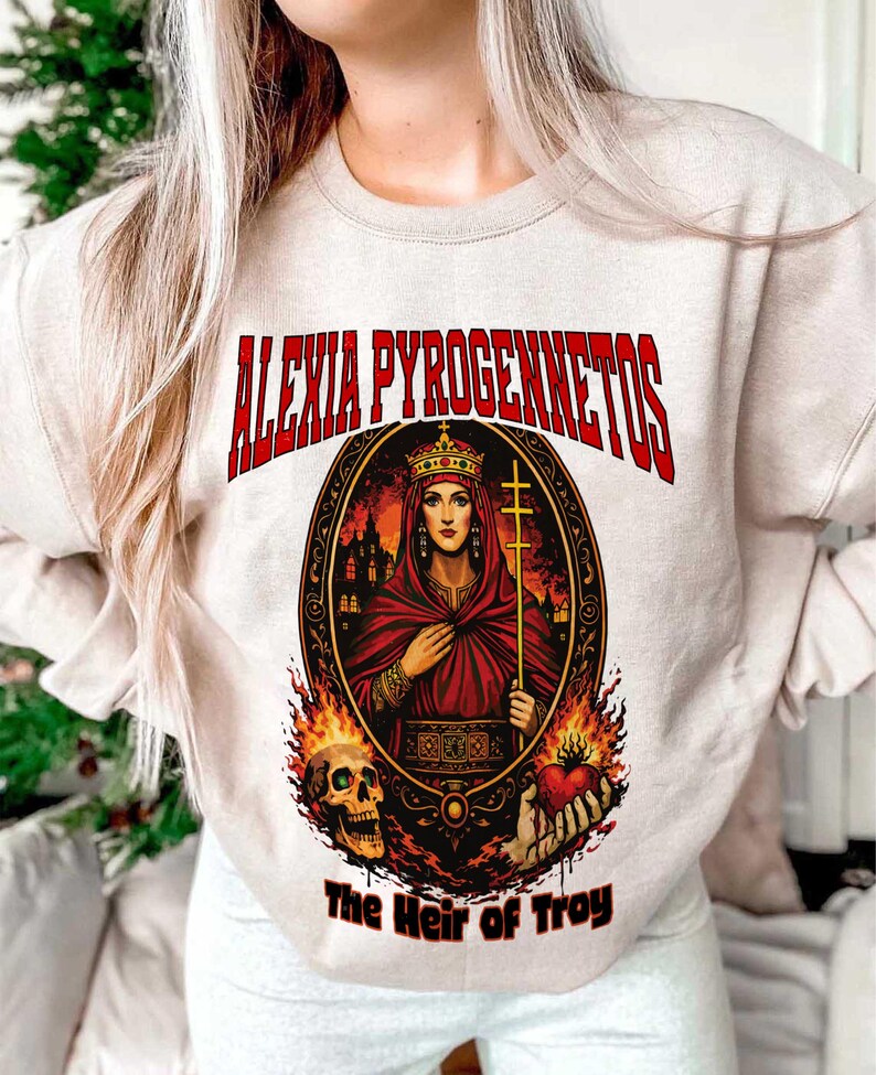 Op de afbeelding: Beige sweatshirt met een afbeelding van een vrouw in een rode mantel en kroon, met een kruis. De tekst "ALEXIA PYROGENNETOS" staat in het rood boven de afbeelding, met "The Heir of Troy" eronder. Vlammen en een schedel zijn ook aanwezig.