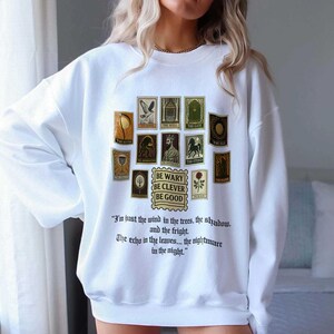 May include: White sweatshirt featuring a collection of vintage-style postage stamp illustrations in shades of brown, green, and gold. The text "BE WARY BE CLEVER BE GOOD" is printed below the stamps, with a quote below.