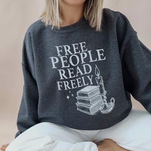 Op de afbeelding: Donkergrijze sweatshirt met de witte tekst "FREE PEOPLE READ FREELY". De afbeelding bevat een stapel boeken, een brandende kaars en sterren. De sweatshirt heeft een casual stijl.