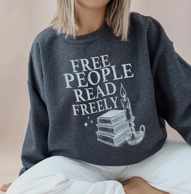 Op de afbeelding: Donkergrijze sweatshirt met de witte tekst "FREE PEOPLE READ FREELY". De afbeelding bevat een stapel boeken, een brandende kaars en sterren. De sweatshirt heeft een casual stijl.