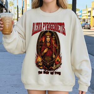 Op de afbeelding: Cr&egrave;mekleurige sweatshirt met een grafisch ontwerp. Het ontwerp bevat de tekst "ALEXIA PYROGENNETOS" in het rood, een portret van een vrouw in een rode jurk, een kroon en een kruis, omringd door vlammen en een schedel. De tekst "The Heir of Troy" staat eronder.