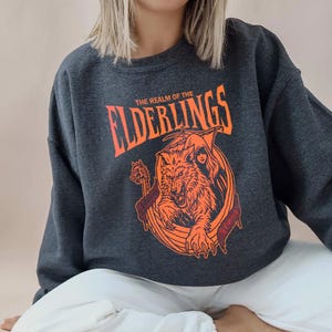 May include: Dark gray sweatshirt with the text "THE REALM OF THE ELDERLINGS" in orange. The graphic features an orange wolf, a hooded figure, and a boat. The sweatshirt is a crew neck style.