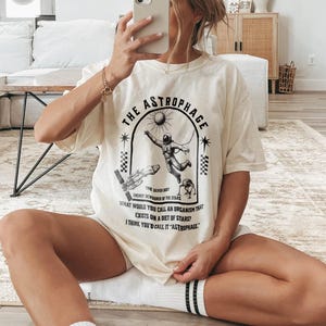 May include: Cream-colored t-shirt with a black graphic design featuring an astronaut, a rocket, and the text "THE ASTROPHAGE". The design includes the phrases "ENERGY DEVOURER OF THE STARS" and "WHAT WOULD YOU CALL AN ORGANISM THAT EXISTS ON A DIET OF STARS?"