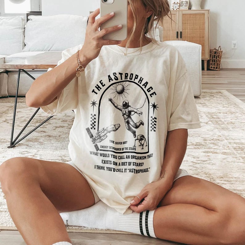 May include: Cream-colored t-shirt with a black graphic design featuring an astronaut, a rocket, and the text "THE ASTROPHAGE". The design includes the phrases "ENERGY DEVOURER OF THE STARS" and "WHAT WOULD YOU CALL AN ORGANISM THAT EXISTS ON A DIET OF STARS?"