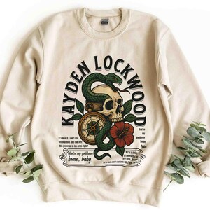 May include: Cream-colored sweatshirt with a graphic design featuring a skull, a green snake, a compass, and a red flower. The text "KAYDEN LOCKWOOD" arches above the design, with additional text below. A casual, unisex garment.