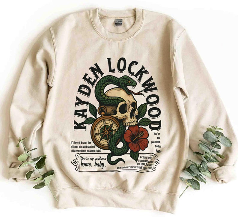 May include: Cream-colored sweatshirt with a graphic design featuring a skull, a green snake, a compass, and a red flower. The text "KAYDEN LOCKWOOD" arches above the design, with additional text below. A casual, unisex garment.