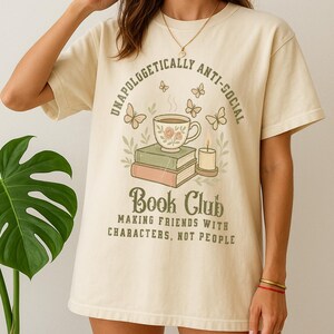 May include: Cream-colored t-shirt with the text "UNAPOLOGETICALLY ANTI-SOCIAL" arched above a graphic of a teacup, books, and a candle. Below, the shirt reads "Book Club: Making Friends with Characters, Not People."