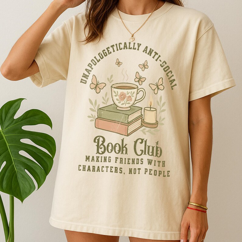 May include: Cream-colored t-shirt with the text "UNAPOLOGETICALLY ANTI-SOCIAL" arched above a graphic of a teacup, books, and a candle. Below, the shirt reads "Book Club: Making Friends with Characters, Not People."