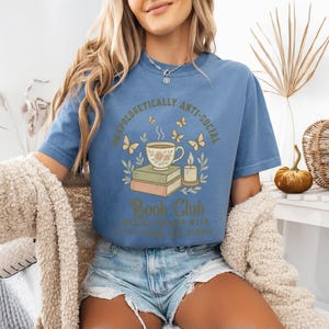 May include: A blue t-shirt with the words "Unapologetically Anti-Social Book Club" printed on the front. The graphic includes a teacup, books, butterflies, and a candle. The shirt is a comfortable, casual style.