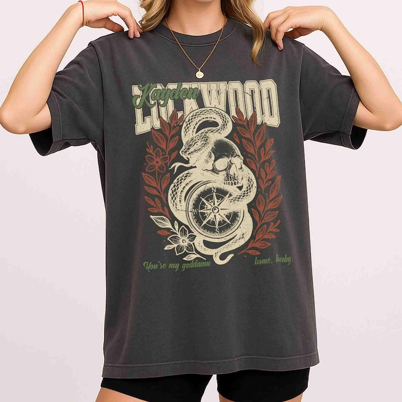 Puede incluir: Camiseta gris oscuro con un dise&ntilde;o gr&aacute;fico que presenta una calavera, una serpiente y una br&uacute;jula. El texto "Lockwood" est&aacute; en la parte superior, con texto adicional en la parte inferior. El dise&ntilde;o incluye elementos florales rojos y verdes.