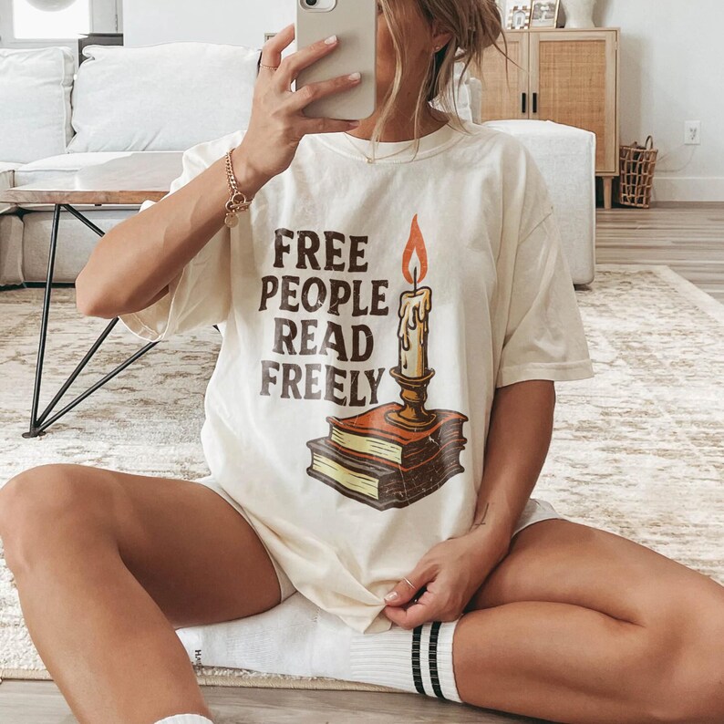 Może przedstawiać: Kremowy t-shirt z napisem "FREE PEOPLE READ FREELY" w brązowych literach. Grafika przedstawia świecę na dw&oacute;ch książkach z pomarańczowym płomieniem. Koszulka ma luźny, obszerny kr&oacute;j.