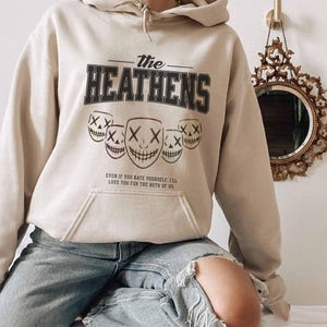 May include: A beige hoodie featuring the text "The Heathens" in black and grey, and five smiling mask graphics. The hoodie also has the quote: "Even if you hate yourself, I'll love you for the both of us."