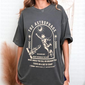 May include: Dark grey t-shirt with a graphic featuring an astronaut, spaceship, and the text "THE ASTROPHAGE". The design includes a sun and stars, with additional text about organisms that eat stars. A casual, oversized fit.