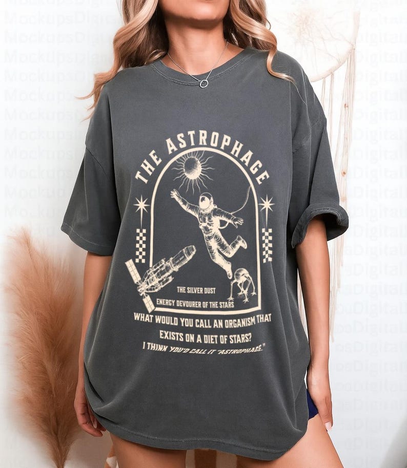 May include: Dark grey t-shirt with a graphic featuring an astronaut, spaceship, and the text "THE ASTROPHAGE". The design includes a sun and stars, with additional text about organisms that eat stars. A casual, oversized fit.