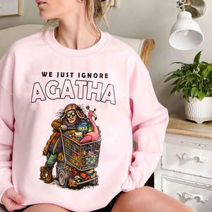 May include: A light pink sweatshirt with the text "WE JUST IGNORE AGATHA" above a cartoon illustration of a person pushing a shopping cart filled with items. The cart includes a pink flamingo and an IKEA box.