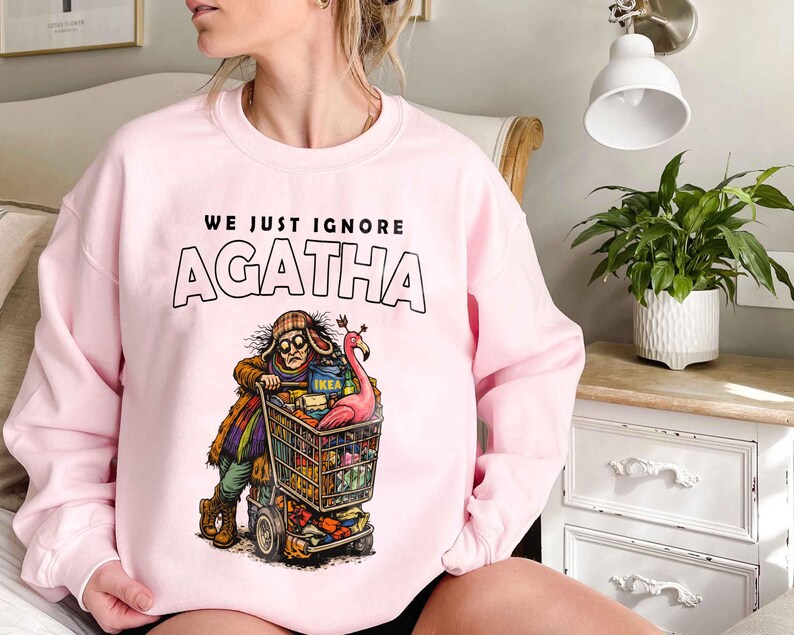 May include: A light pink sweatshirt with the text "WE JUST IGNORE AGATHA" above a cartoon illustration of a person pushing a shopping cart filled with items. The cart includes a pink flamingo and an IKEA box.