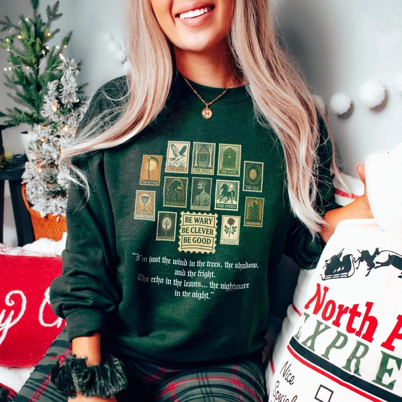 May include: Dark green sweatshirt featuring a collection of vintage-style postage stamp designs in gold. The sweatshirt includes the phrases "BE WARY BE CLEVER BE GOOD" and a quote. The image is of a person wearing the sweatshirt.