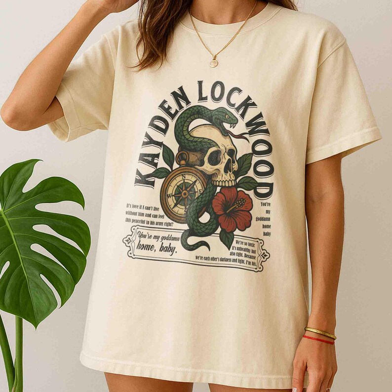 May include: Cream-colored t-shirt featuring a graphic design with the text "KAYDEN LOCKWOOD". The design includes a skull, a green snake, a compass, and a red hibiscus flower. Additional text is printed below the main design.