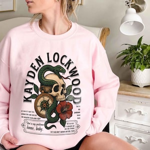 May include: A light pink sweatshirt with the text "KAYDEN LOCKWOOD" in a curved arch. The graphic includes a skull, a green snake, a compass, and a red flower. Additional text is below the graphic.